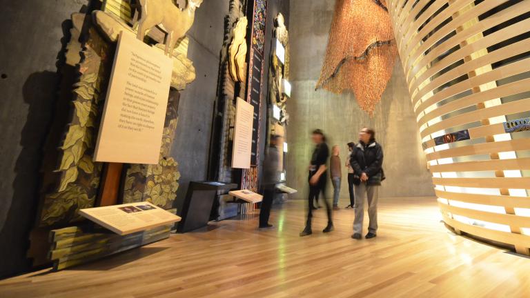 Five museum visitors look at towering exhibits in the “Indigenous Perspectives” gallery at the Canadian Museum for Human Rights. The nearest is made of wood and features trees, animals and a plaque. Behind the visitors, a rounded theatre built of bent wooden slats is visible. Partially obscured.
