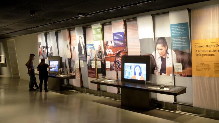 Two people interacting with one of two video screens in a museum gallery. There are images of people and coloured text blocks on the wall.