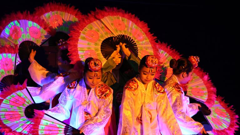 Korean dancers holding large, flower-patterned fans are grouped together so their fans form a circle.