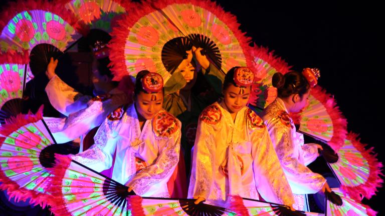 Korean dancers holding large, flower-patterned fans are grouped together so their fans form a circle. Partially obscured.