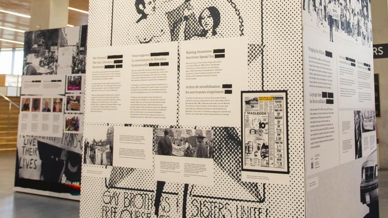 Exhibit in a modern space showcasing historical posters and text about 2SLGBTQ+ activism, with people observing the display and a person on a balcony above.