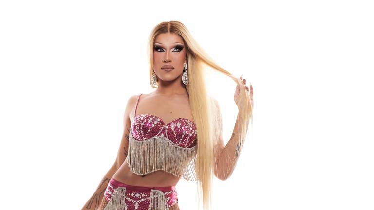 A glamourous drag queen with long blonde hair and dramatic makeup is wearing a bejeweled red bikini top with long silver fringe. With their right hand to their head, a tattoo is revealed on the inner arm that reads “life is blind.” Partially obscured.