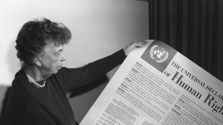 A person woman-presenting holds a large piece of paper covered with text and a large title reading "The Universal Declaration of Human Rights." Partially obscured.