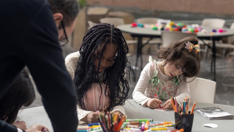 Two children sit at a table making art while an adult leans on the table, watching. Partially obscured.
