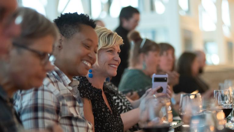 A group of people sit at a long dining table, laughing and enjoying a meal together. The focus is on two people smiling brightly - one wearing a plaid shirt and the other in a black patterned top, with glasses and plates arranged neatly in front of them. Partially obscured.