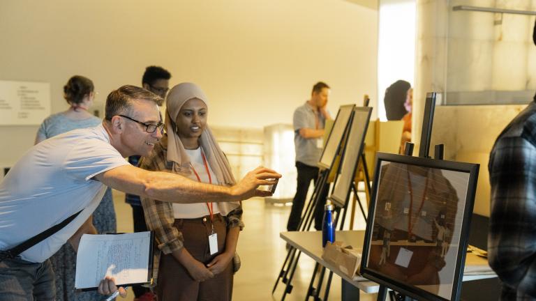 Students in a museum gallery display their work on easels. The focus is on one student talking with an adult who is smiling and taking a photograph with their phone of the student’s work. Partially obscured.