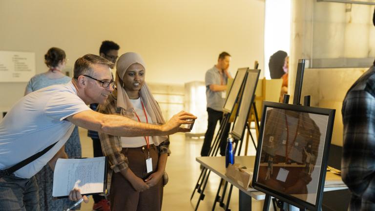 Students in a museum gallery display their work on easels. The focus is on one student talking with an adult who is smiling and taking a photograph with their phone of the student’s work. Partially obscured.
