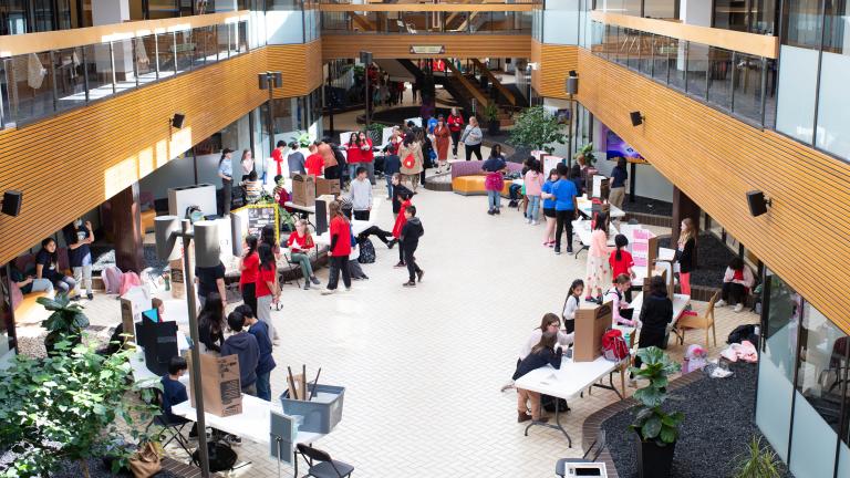 Many people gathered in an open indoor space with tables and posters set up on them. Partially obscured.