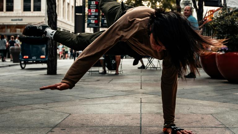 A woman in a grey jacket and darker grey pants is breakdancing on a sidewalk during daytime. Trees and buildings are in the background. Partially obscured.
