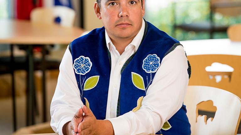 A man wearing a blue vest with flower beadwork, seated with his hands resting on a table. Partially obscured.