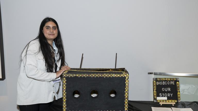 A young woman stands next to a box with an ornamental painted trim. The box has three holes that give a view into the box. 