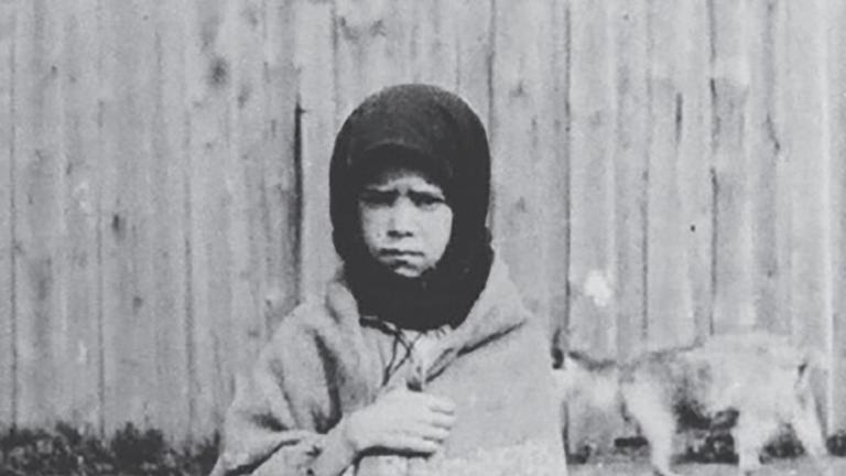 A young girl stands facing the camera with a sorrowful expression, clutching a shawl around her. Partially obscured.