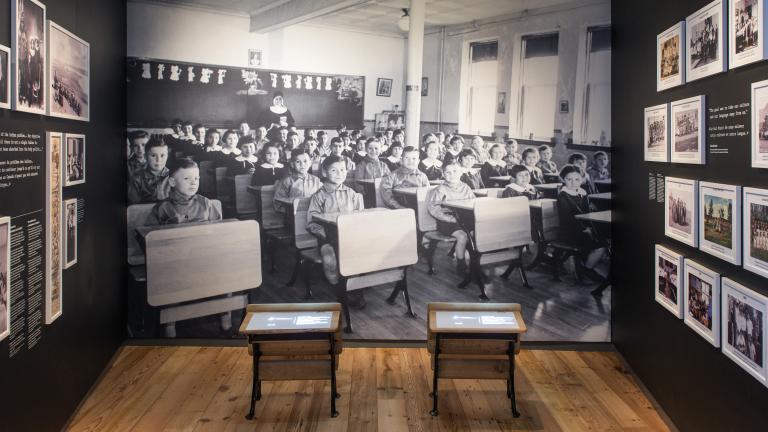 A museum exhibit that includes small school desks in front of a large photographic background of Indigenous children sitting at their desks in a classroom. Partially obscured.