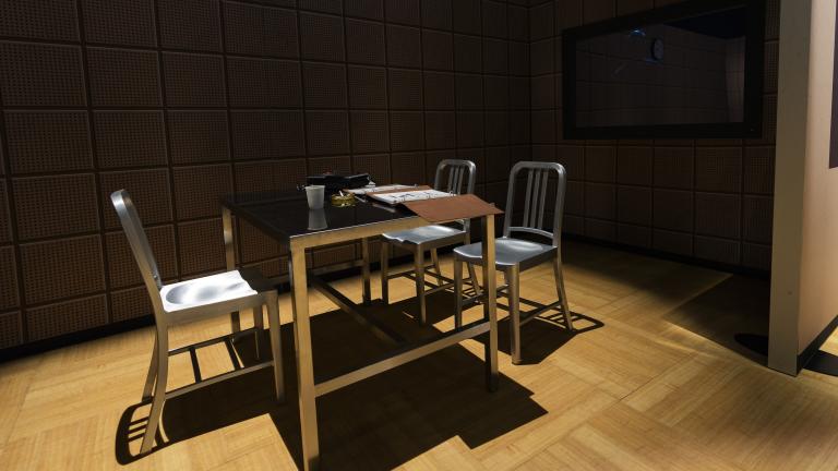 Three metal chairs and a metal table covered with binders and a tape recorder are in the middle of a darkened space. A clock hangs on the wall above the table. A mirrored surface is to the right of the table.