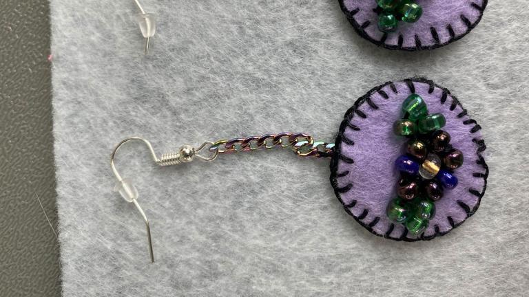 A pair of light purple felt earrings with dark purple sewn borders. In the middle of the earrings are beaded flowers. Partially obscured.