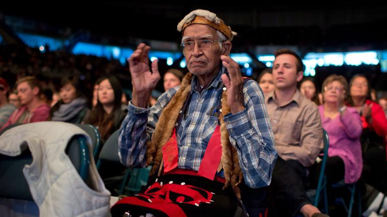 An elderly man wearing a checkered shirt and traditional Indigenous accessories is sitting in a crown. His arms are bent at the elbows and his hands are up facing each other.