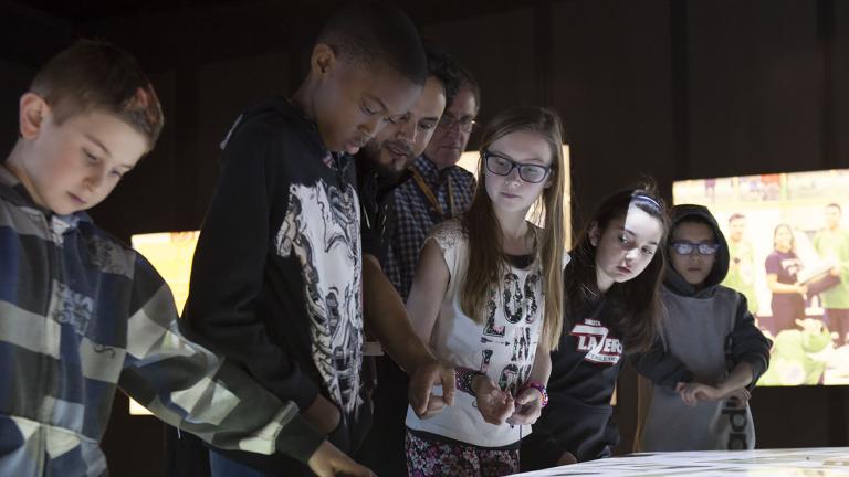 A Museum guide showing five students how to engage with the digital table, around which they are all standing. Partially obscured.