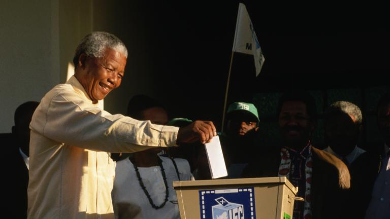 Nelson Mandela extending his right arm to deposit a folded paper in a ballot box. A few people are standing around him.