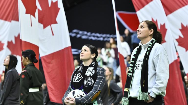 People stand on a stadium field during a pre-game ceremony as large Canadian flags are held behind them, filling the background with red and white. Partially obscured.