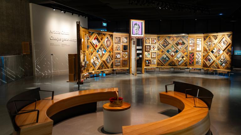 A museum exhibition featuring a large, curved freestanding wooden wall on which is mounted numerous small objects arranged in diamond and rectangular patterns. In the foreground, there circular benches creating an inviting space for visitors to view and contemplate the exhibit.