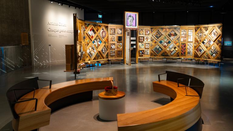 A museum exhibition featuring a large, curved freestanding wooden wall on which is mounted numerous small objects arranged in diamond and rectangular patterns. In the foreground, there circular benches creating an inviting space for visitors to view and contemplate the exhibit. Partially obscured.