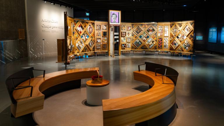 A museum exhibition featuring a large, curved freestanding wooden wall on which is mounted numerous small objects arranged in diamond and rectangular patterns. In the foreground, there circular benches creating an inviting space for visitors to view and contemplate the exhibit. Partially obscured.