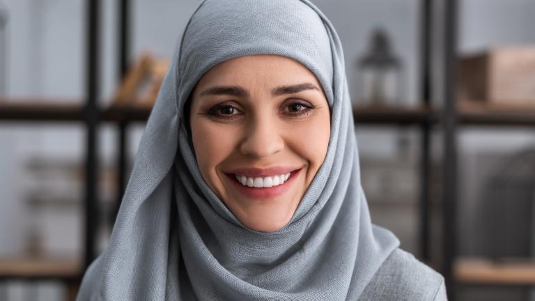 A woman wearing a blue hijab smiles toward the camera, with a bookshelf in the background. Partially obscured.