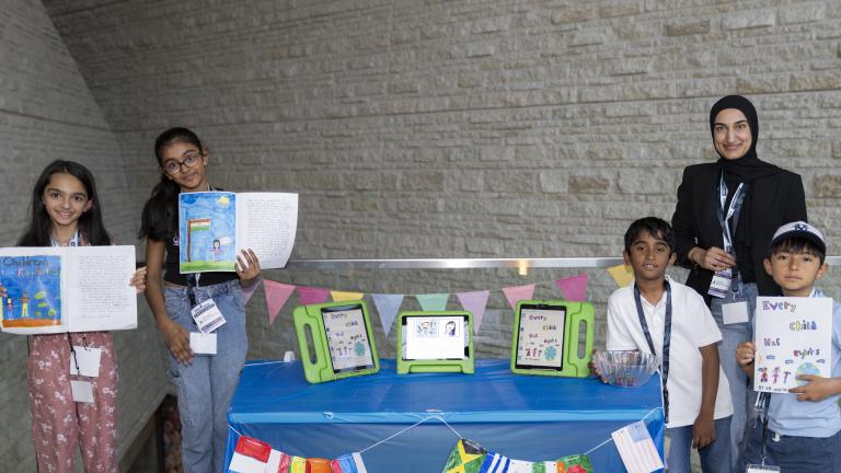 Four students and an adult stand next to a table with flags from countries around the world. Three of them are holding up an open book.
