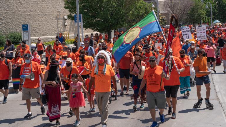 Hundreds of demonstrators in orange shirts, many also wearing traditional Indigenous articles such as a headdress or ribbon skirt and carrying signs, march down a wide paved street. Partially obscured.