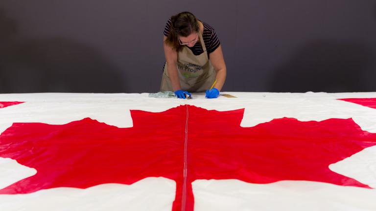 A woman wearing protective gloves and an apron takes notes behind a Canadian flag, which lies flat on a table in the foreground.