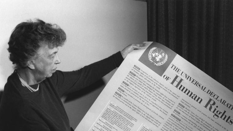 A person holds a large piece of paper covered with text and a large title reading "The Universal Declaration of Human Rights." Partially obscured.