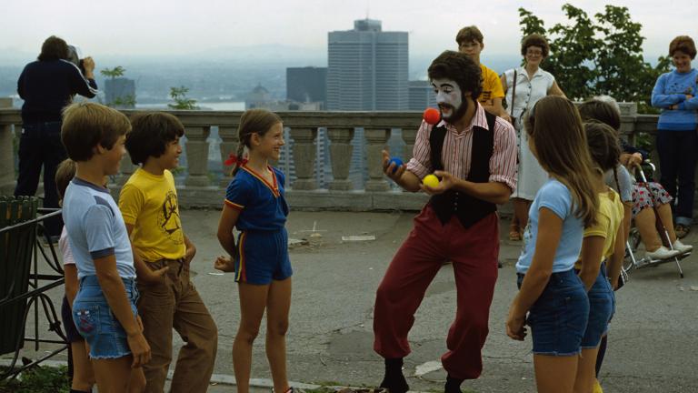 A man with a painted face juggles outdoors, as smiling children watch. A city skyline appears behind him at a distance. Partially obscured.