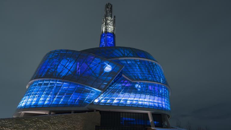 The exterior of the Canadian Museum for Human Rights is pictured at night. The building is lit up from the inside with vibrant deep blue lights. Partially obscured.