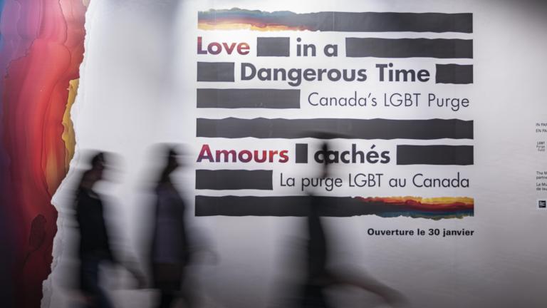 Three silhouettes of people walking past a large wall panel featuring the logo for the Love in a Dangerous Time exhibition. The silhouettes are blurred as the individuals are walking from left to right. Partially obscured.