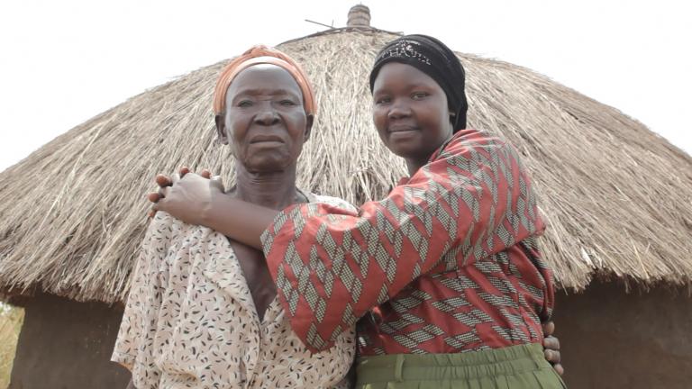 Two women are standing in front of a hut with a straw roof, looking at the camera. The younger woman on the right is smiling and embracing the one on the left. Partially obscured.