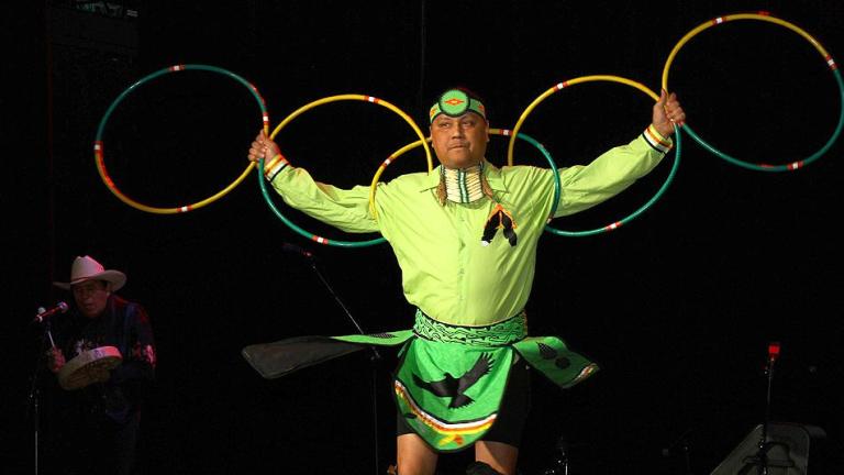 A hoop dancer dressed in a long-sleeve yellow top and green skirt is holding five hoops with arms outstretched like an eagle.