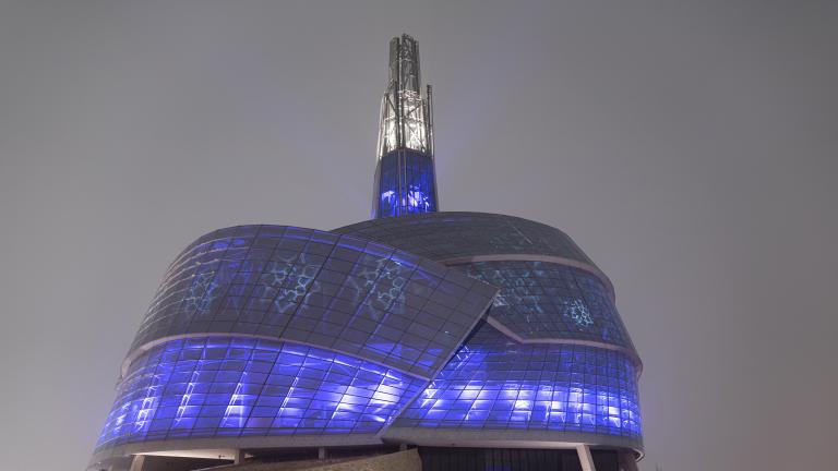 A building surrounded by glass panes illuminated in blue and purple. Snowflake designs are also projected on the windows. Partially obscured.