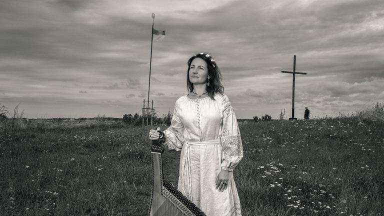 A woman in a long white dress stands in a field holding a bandura, a Ukrainian plucked string folk instrument. Partially obscured.
