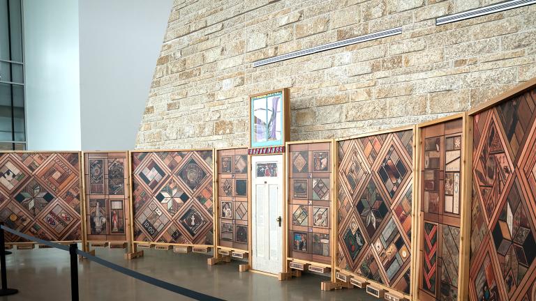 Right view of a structure of vertical wood panels covered in patterns of images, with a white wooden door at the centre. Partially obscured.