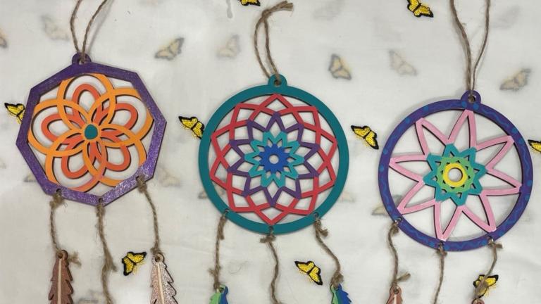 Three colourful dream catchers made from painted wood lie on butterfly-festooned craft paper. Partially obscured.