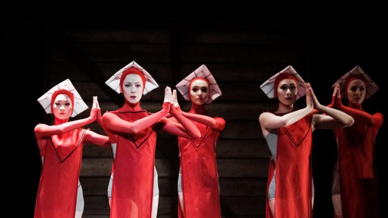 On a dark stage are five ballet dancers dressed in red knee-length flowy costumes with white square-like hats. They are looking forward with their hands in prayer. Partially obscured.
