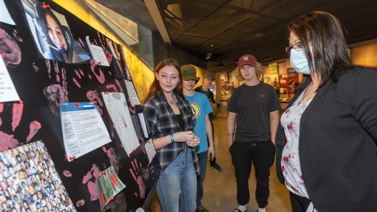 Three students stand in front of a poster board, placed on a table. A museum visitor stands in front of them looking at the poster board. Partially obscured.