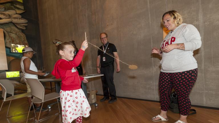 A young girl holds a small stick attached to a string and a small circular piece of cardboard. She is swinging this rope and cardboard as two adults watch. Partially obscured.
