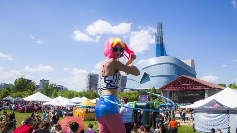 A person in brightly coloured wig and clothing swinging a hula hoop at an outdoor festival, with the Museum in the background. Partially obscured.