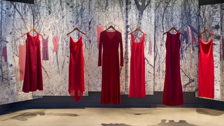 Five red dresses, representing missing and murdered Indigenous women and girls, hang in a row in the Canadian Journeys gallery as part of an exhibit titled “From Sorrow to Strength.” Behind the dresses, the scene of a snow-covered forest with more red dresses hung on the branches of trees is displayed on a series of banners. Partially obscured.