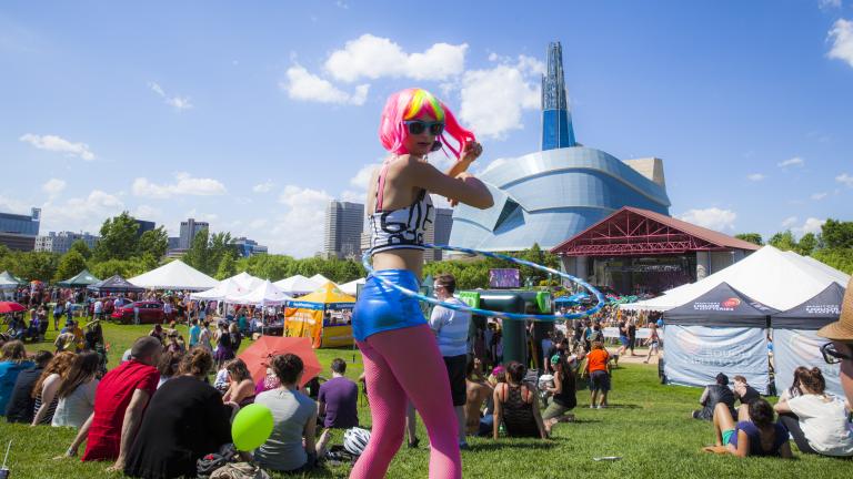 A hula hoop dancer with a pink and fluorescent yellow wig with pink leggings walks in a crowd of people sitting on a field of grass in front of a large glass building. Partially obscured.