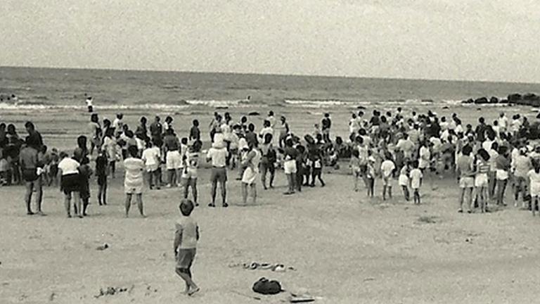 A black-and-white photograph of a crowd of people, most of them standing, on a beach. Partially obscured.
