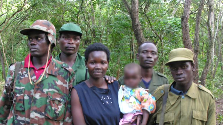 A woman is holding a baby, while four men in army fatigues stand beside and behind her. They are all standing in front of a forest, posed for the camera. Partially obscured.
