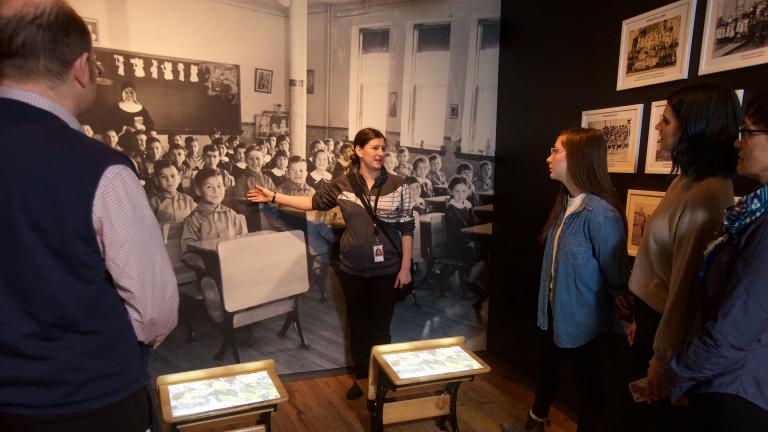 A visitor looks at a gallery niche on residential schools at the Canadian Museum for Human Rights. Two small desks sit in the middle of the niche. A large image of Indigenous students sitting at desks at a residential school is on the back wall of the niche, with images and artifacts on the side walls. Partially obscured.