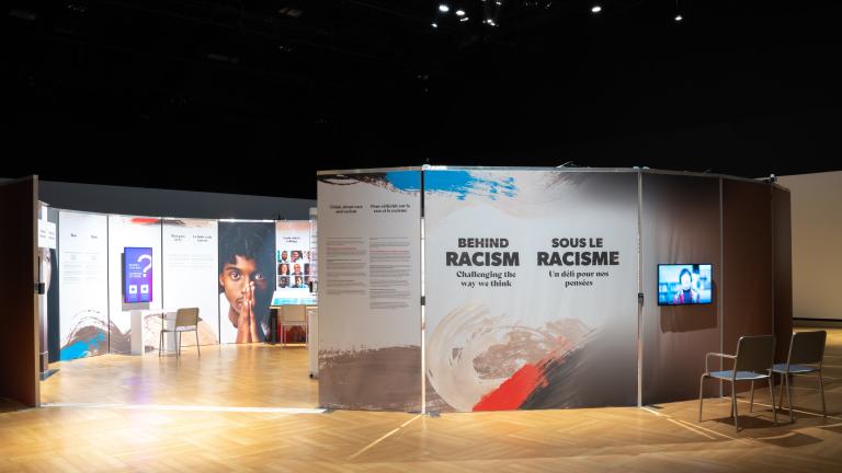 Upright panels are arranged in a circle in a dimly lit room. A gap offers people a way to enter the circle, and the interior of the circle is lit brightly. Next to the opening, a panel shows the words "Behind Racism: Challenging the Way We Think." Partially obscured.